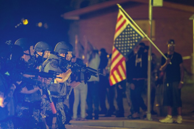 A police officer raises his weapon at a car speeding in his general direction as a more vocal and confrontational group of demonstrators stands on the sidewalk during further protests in reaction to the shooting of Michael Brown near Ferguson, Missouri August 18, 2014. Police fired tear gas and stun grenades at protesters in Ferguson, Missouri on Monday, after days of unrest sparked by the fatal shooting of an unarmed black teenager by a white policeman. REUTERS/Lucas Jackson